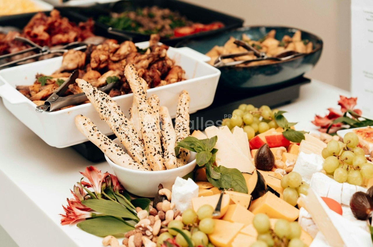 Plateau de fromages, fruits, noix et gressins avec des plats de viande et légumes en arrière-plan sur une table blanche.