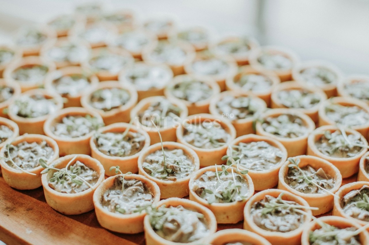 Petites tartelettes garnies de crème et décorées de jeunes pousses, disposées en rangées sur une table en bois.