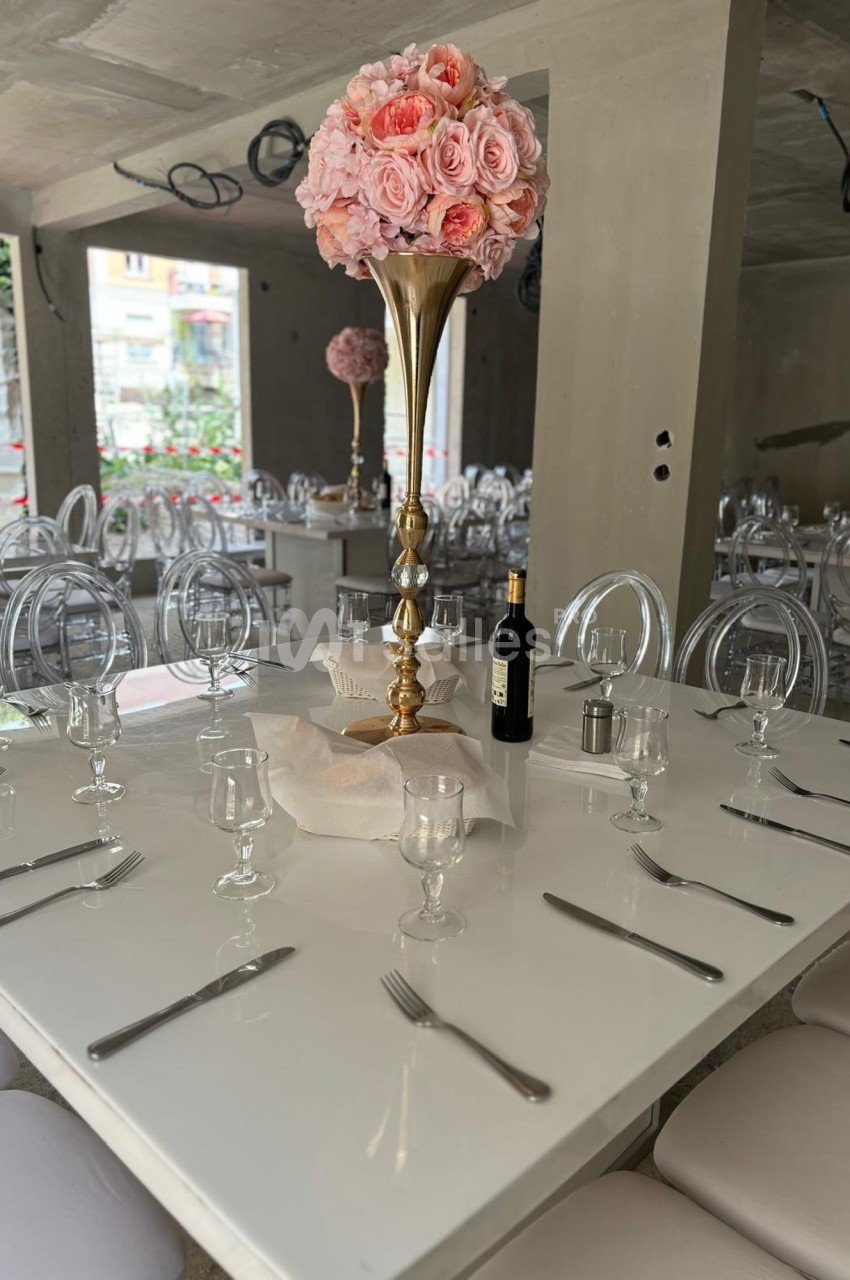 Table dressée avec nappes blanches, couverts, verres, centre de table floral rose et bouteille de vin dans une salle moderne.