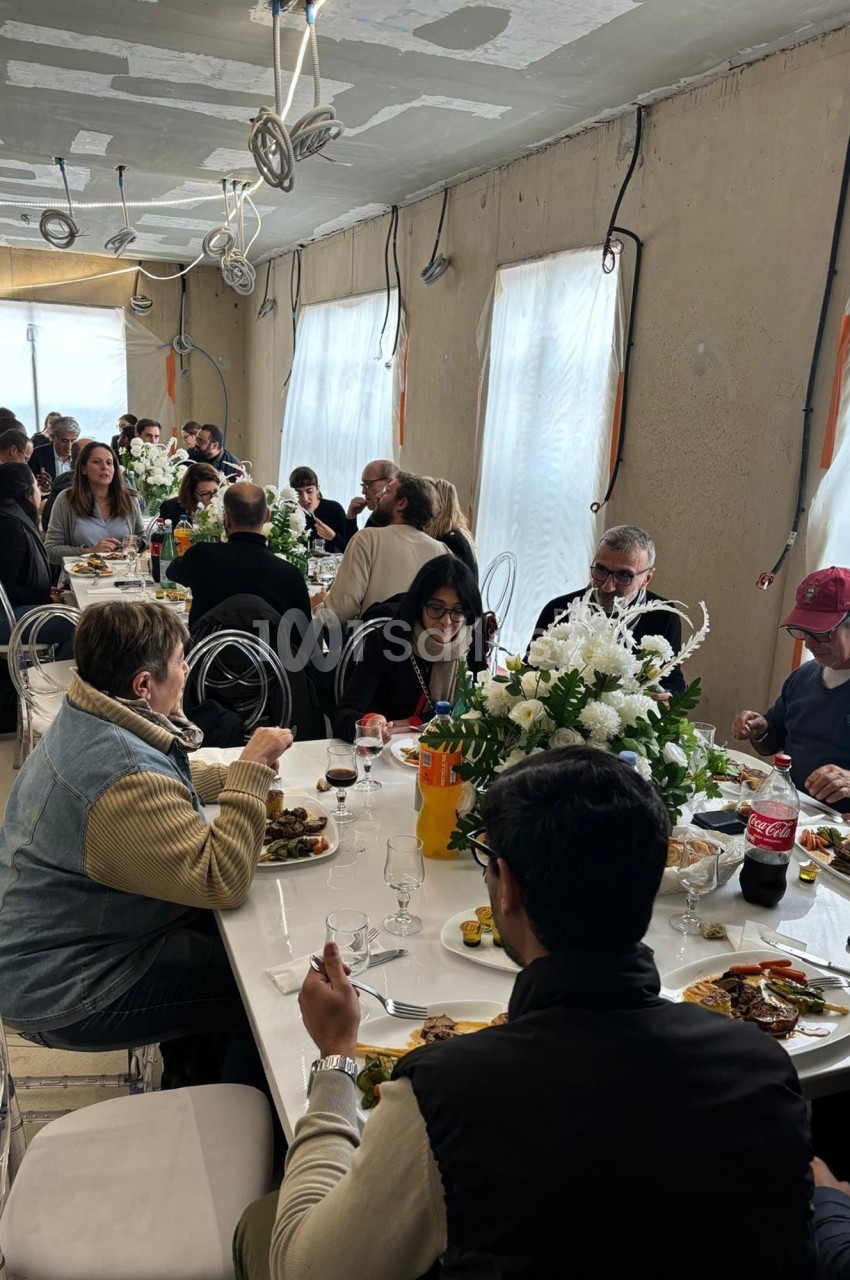 Des personnes partagent un repas convivial autour de grandes tables décorées de fleurs blanches dans une salle lumineuse.
