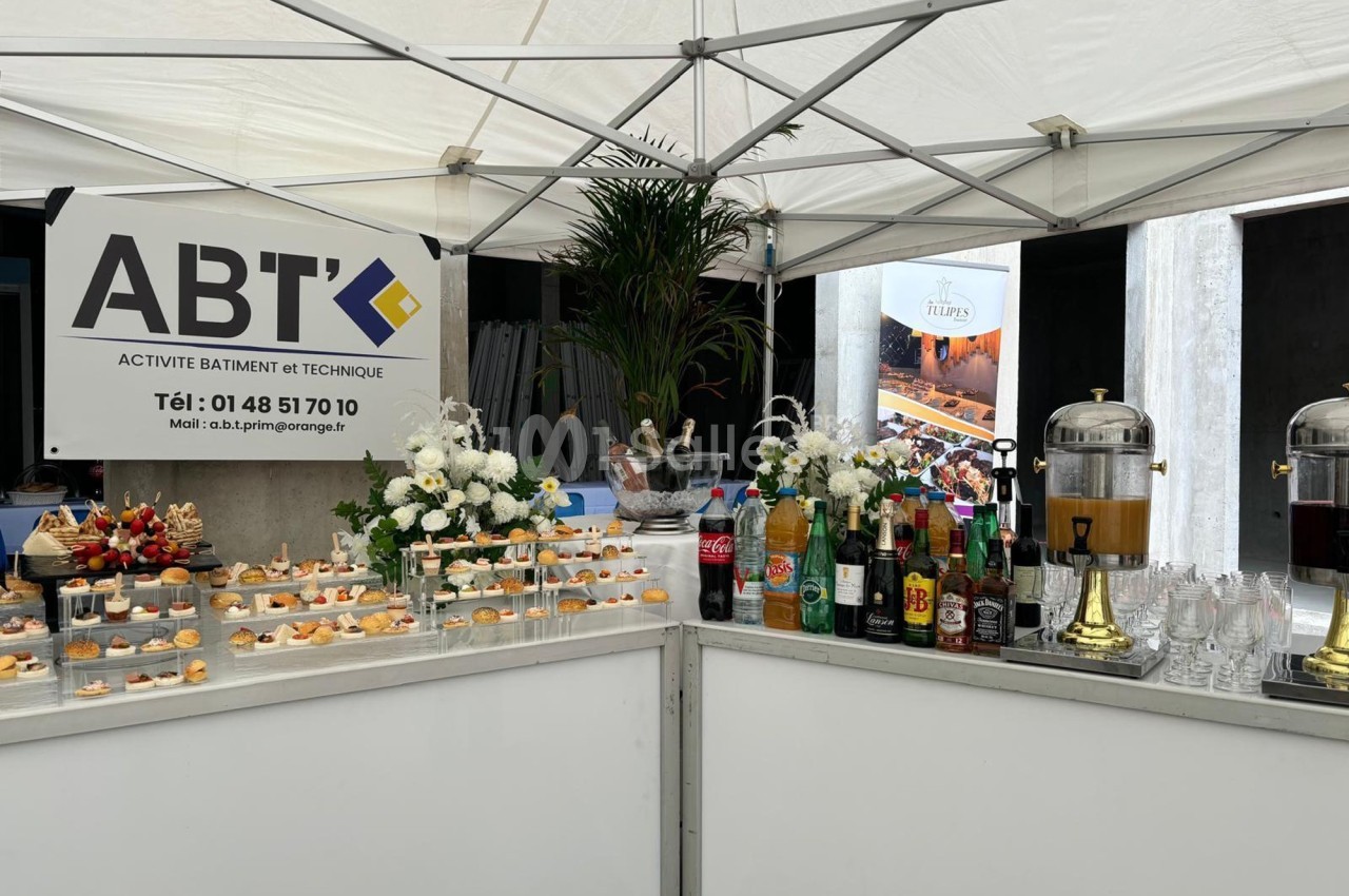 Buffet sous une tente avec boissons, amuse-bouches et décoration florale, devant un panneau d'entreprise ABT.