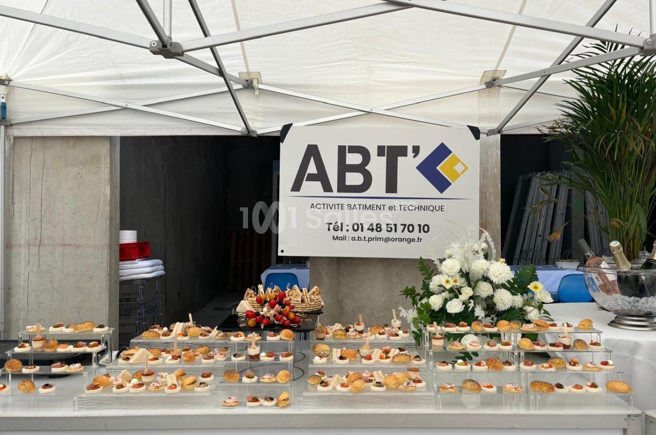 Buffet de petits fours disposé sous une tente blanche, avec un panneau d'entreprise en arrière-plan.