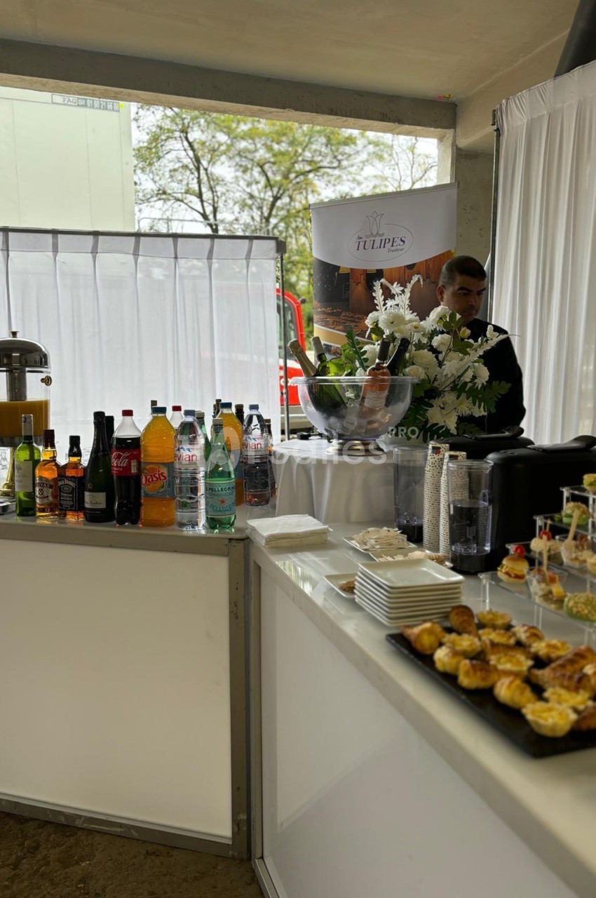 Buffet avec boissons variées, viennoiseries et fleurs blanches, installé dans un espace lumineux avec rideaux blancs.