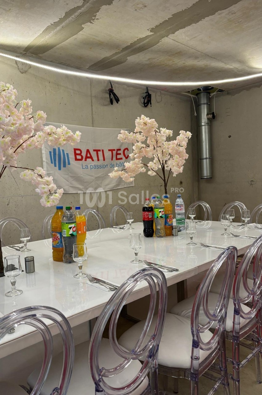Table dressée avec des chaises transparentes, boissons variées et décorations florales dans une salle au mur brut.