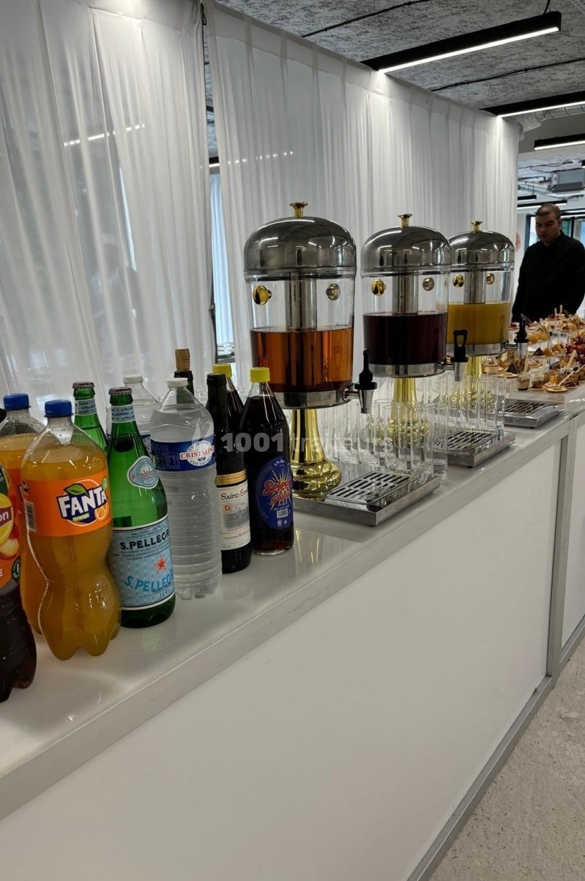 Table avec distributeurs de boissons, bouteilles variées et verres alignés dans un espace lumineux.