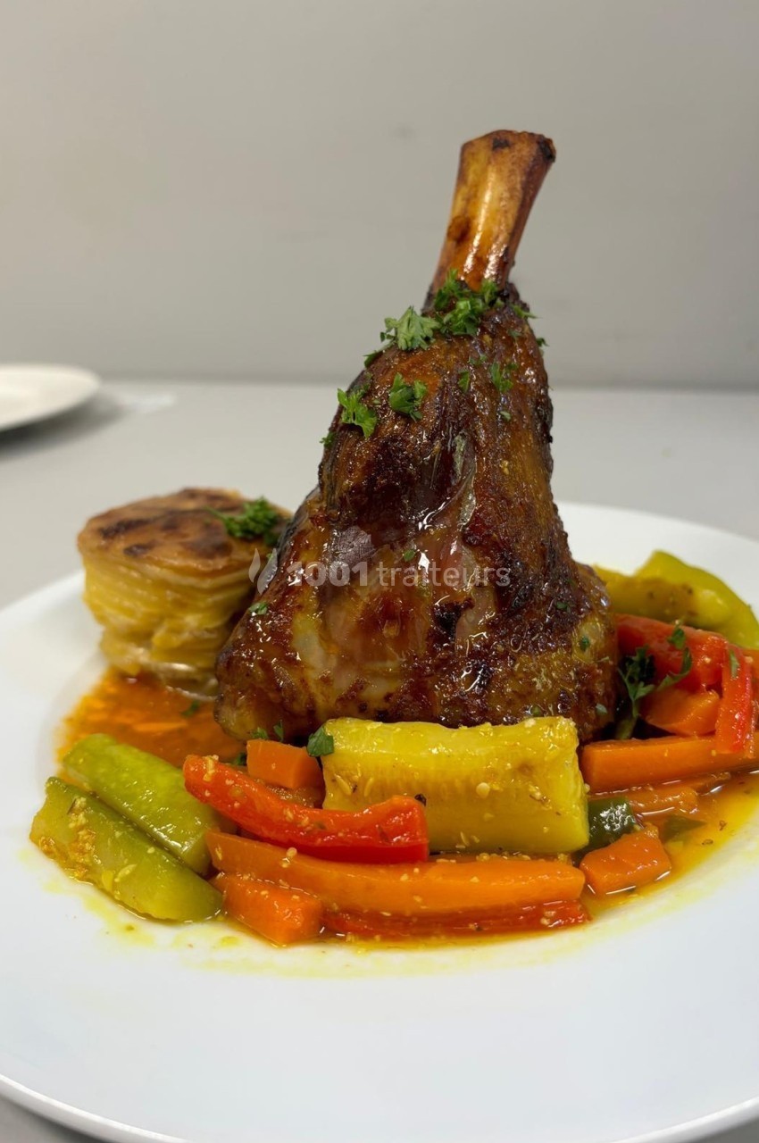 Jarret d'agneau braisé servi avec des légumes colorés et un gratin de pommes de terre, présenté sur une assiette blanche.