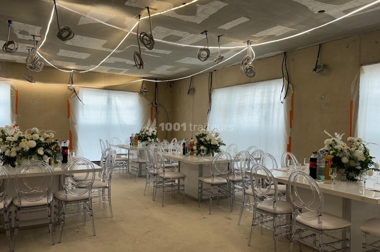 Salle en cours d'aménagement avec des tables décorées de fleurs blanches, chaises transparentes et éclairage suspendu.