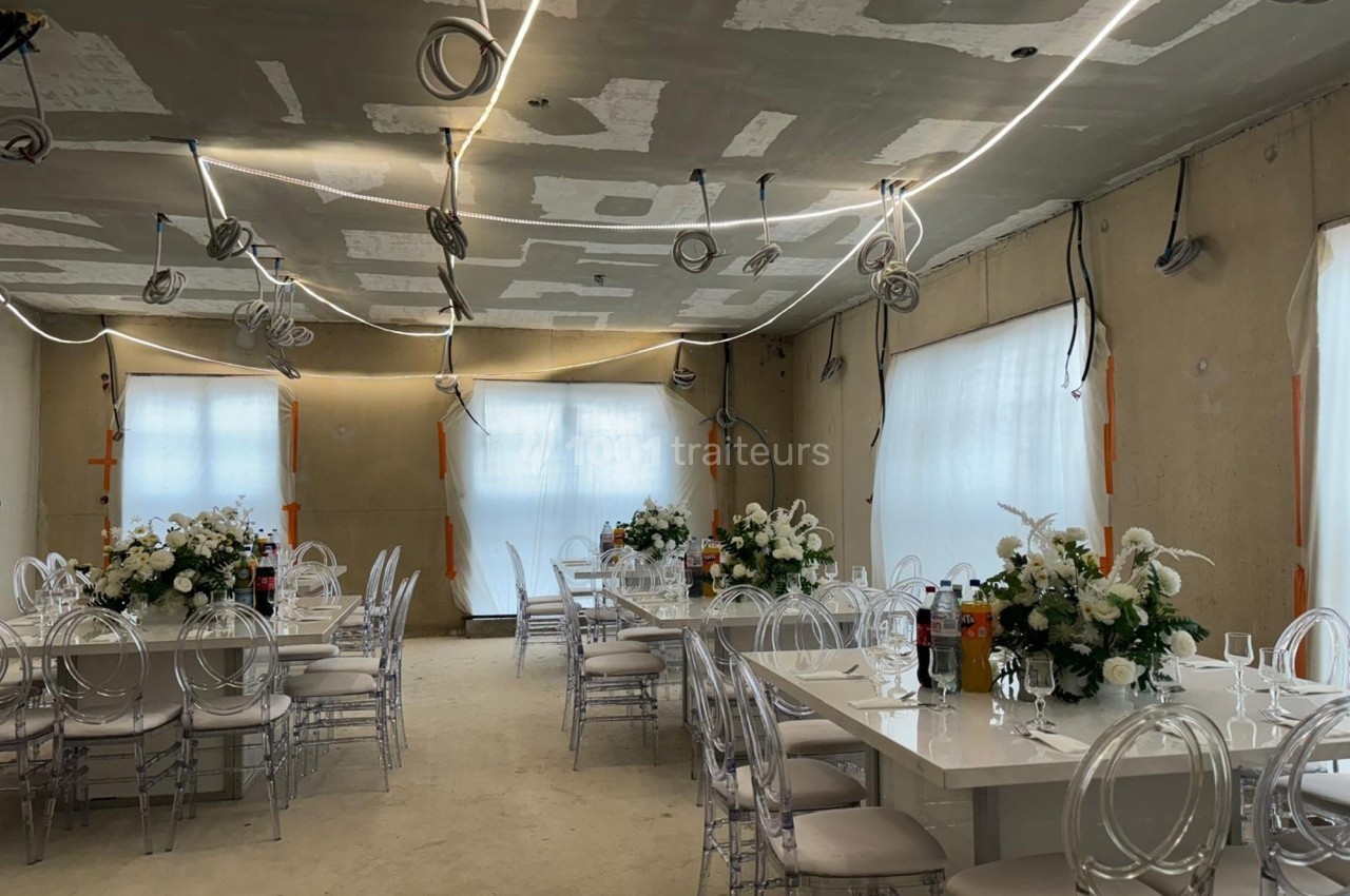 Salle en cours d'aménagement avec des tables décorées de fleurs blanches et des chaises transparentes.