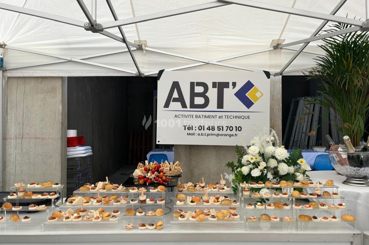 Buffet de réception sous une tente blanche, avec des plateaux de petits fours et une enseigne d'entreprise en arrière-plan.