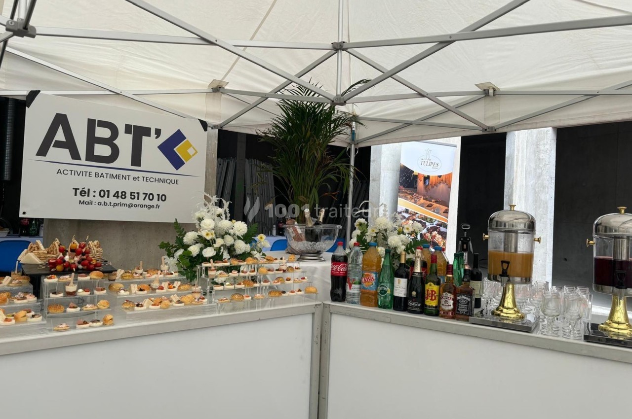 Buffet sous une tente avec boissons, amuse-bouches et décoration florale, accompagné d'un panneau d'entreprise.