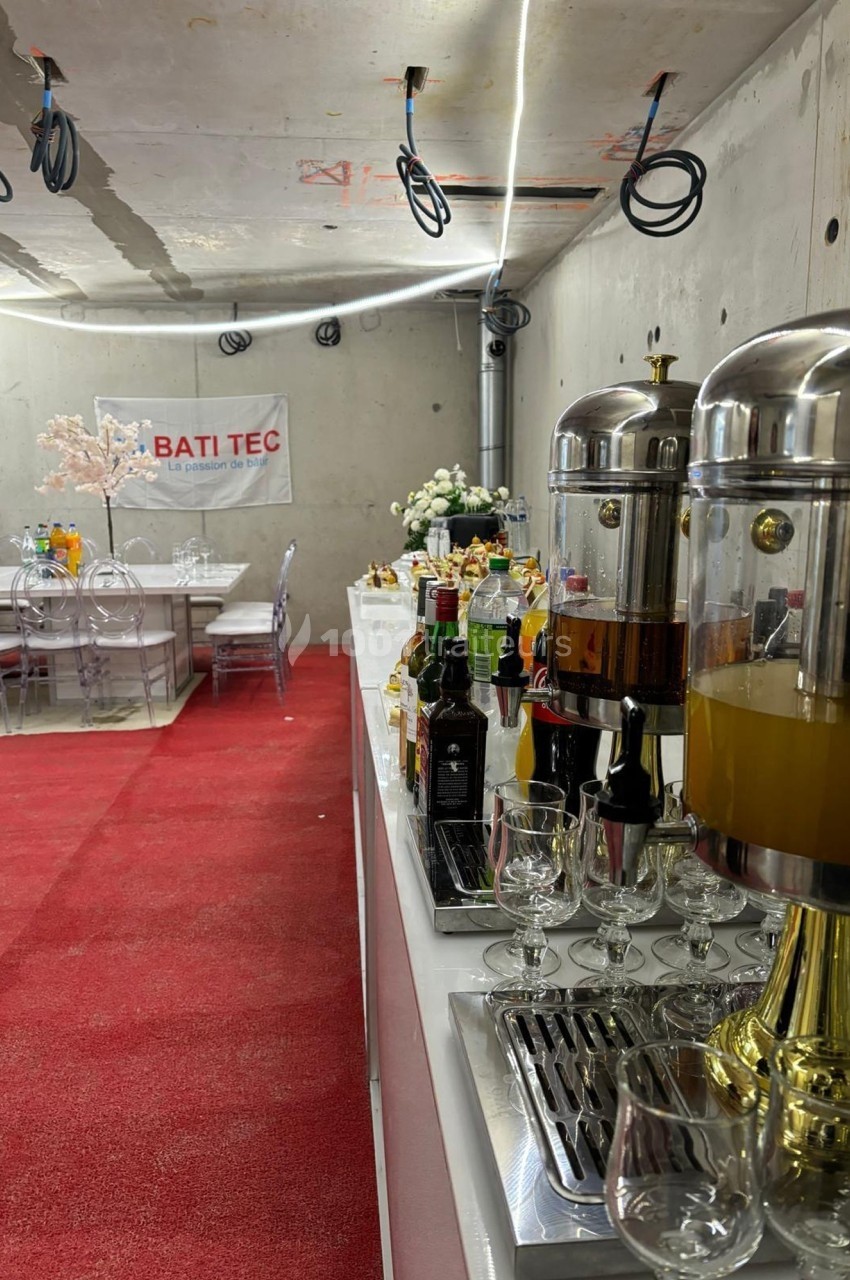 Buffet avec boissons et jus en premier plan, salle aménagée avec tables et chaises transparentes en arrière-plan.
