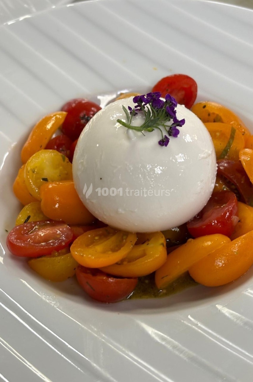 Assiette de burrata garnie de tomates cerises colorées et décorée d'une fleur violette sur une table blanche.