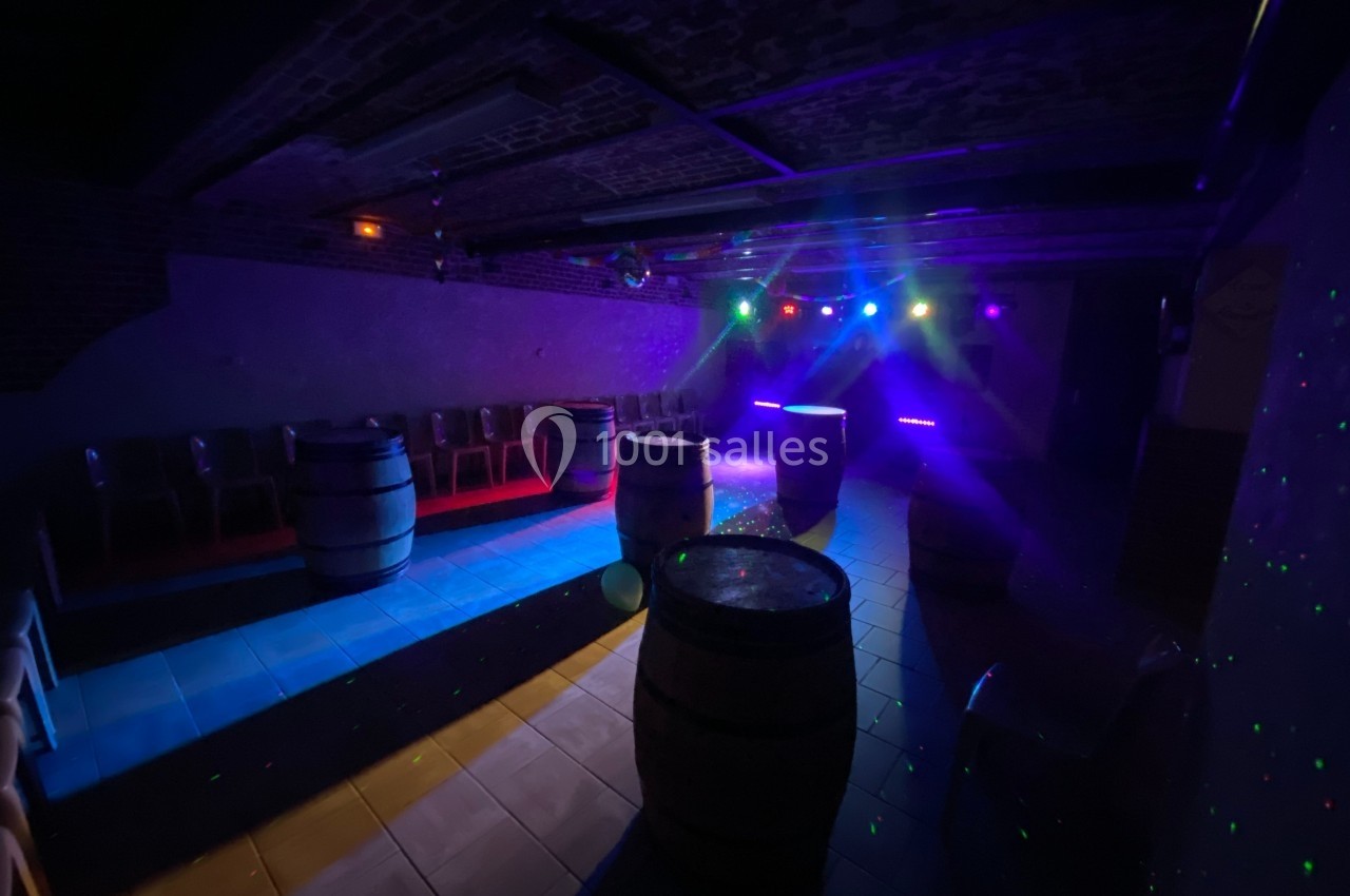 Salle sombre avec tonneaux en bois, chaises alignées contre un mur et éclairage coloré de type discothèque.