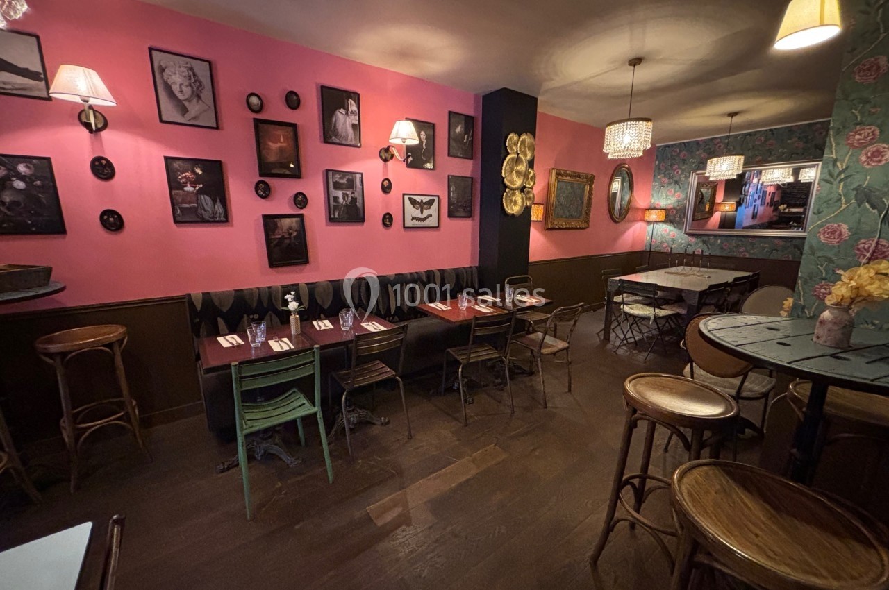 Salle de restaurant cosy avec murs roses décorés de cadres, tables en bois et chaises dépareillées.