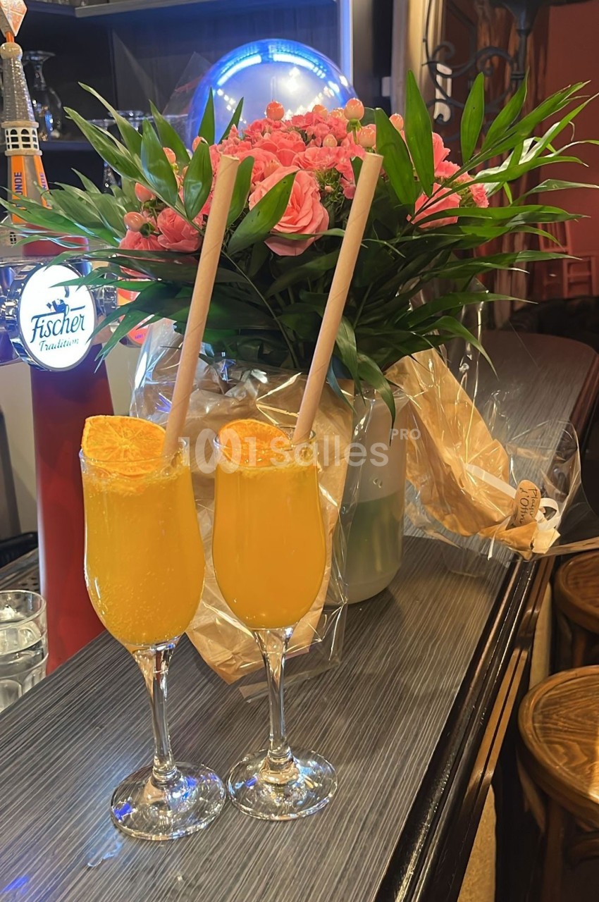 Deux verres de cocktail orange avec des pailles en papier, posés sur un comptoir devant un bouquet de fleurs roses.