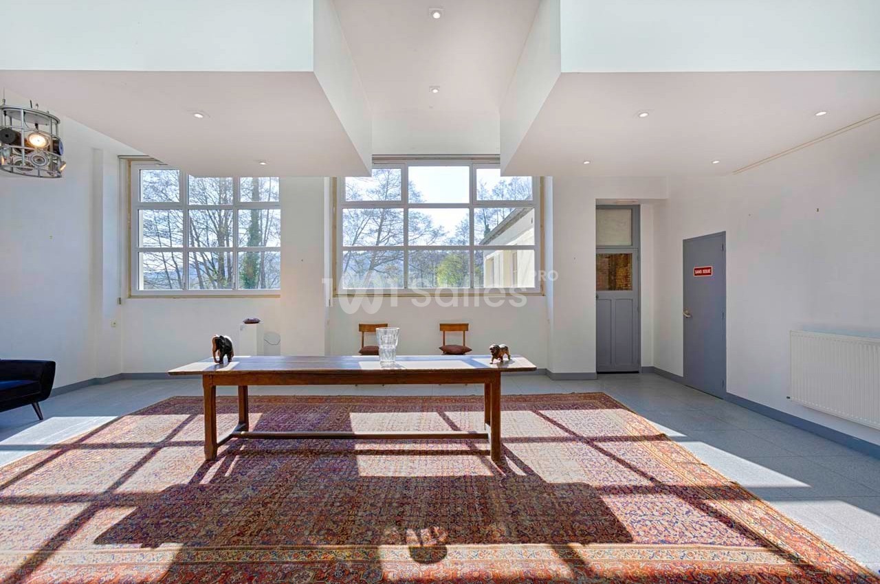 Salle lumineuse avec de grandes fenêtres, une table en bois au centre et un tapis coloré au sol.