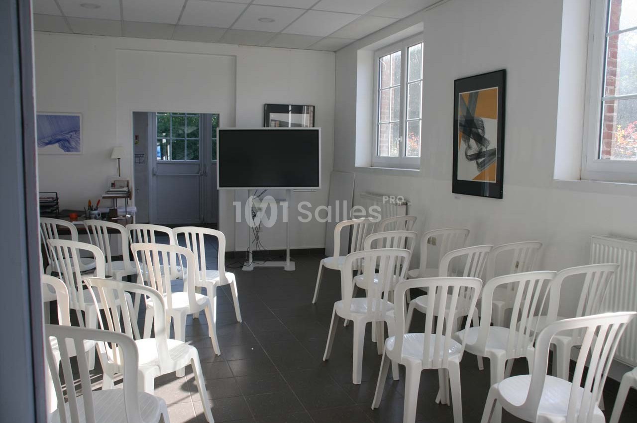 Salle lumineuse avec des chaises blanches disposées face à un écran noir sur pied, près de fenêtres et de tableaux.