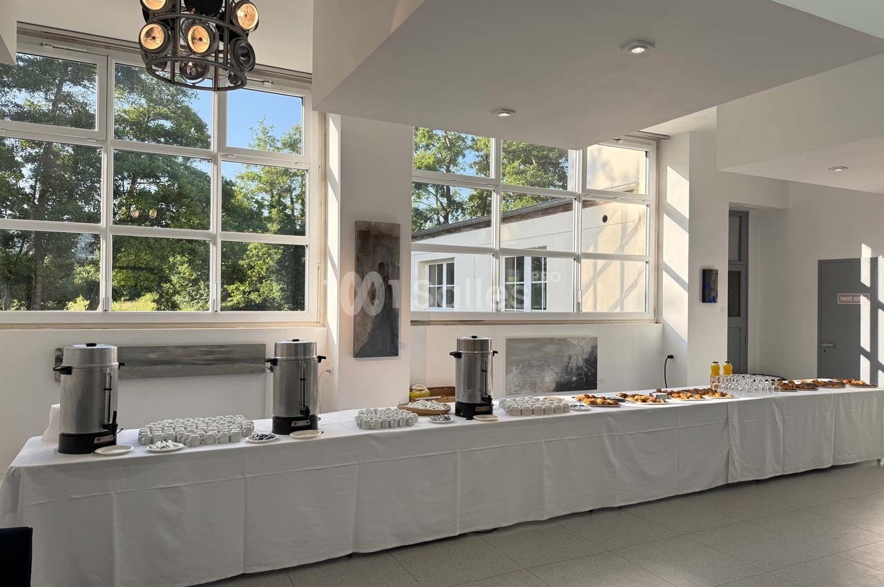 Table dressée avec boissons chaudes, jus et viennoiseries dans une salle lumineuse avec de grandes fenêtres.