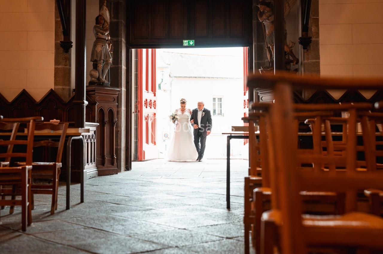 Un couple entre dans une église par une porte ouverte, vu depuis l'intérieur avec des chaises en bois au premier plan.