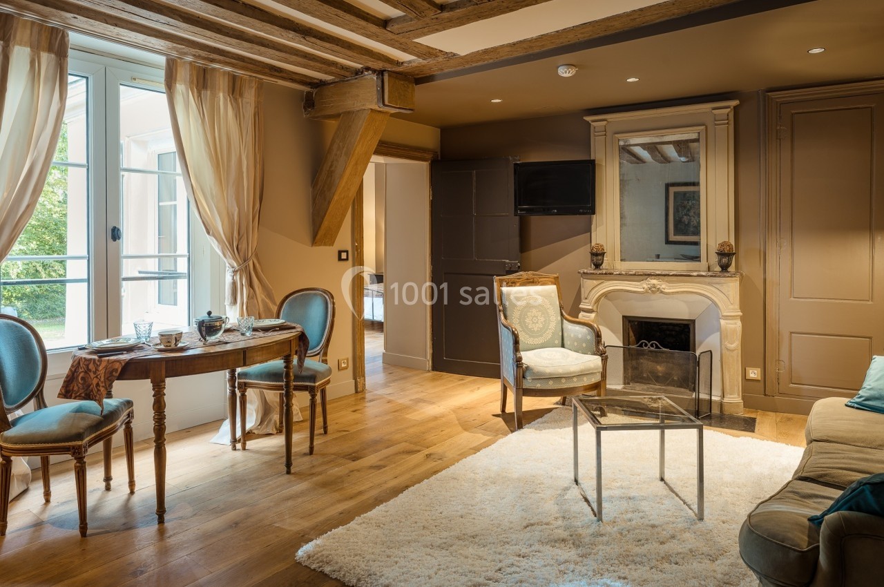 Salon chaleureux avec poutres apparentes, cheminée, table dressée, fauteuils et grande fenêtre donnant sur l'extérieur.