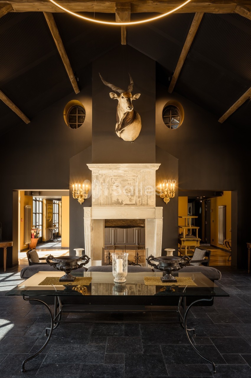 Salle de séjour sombre avec cheminée en pierre, tête de cerf au mur, table en verre et poutres apparentes au plafond.