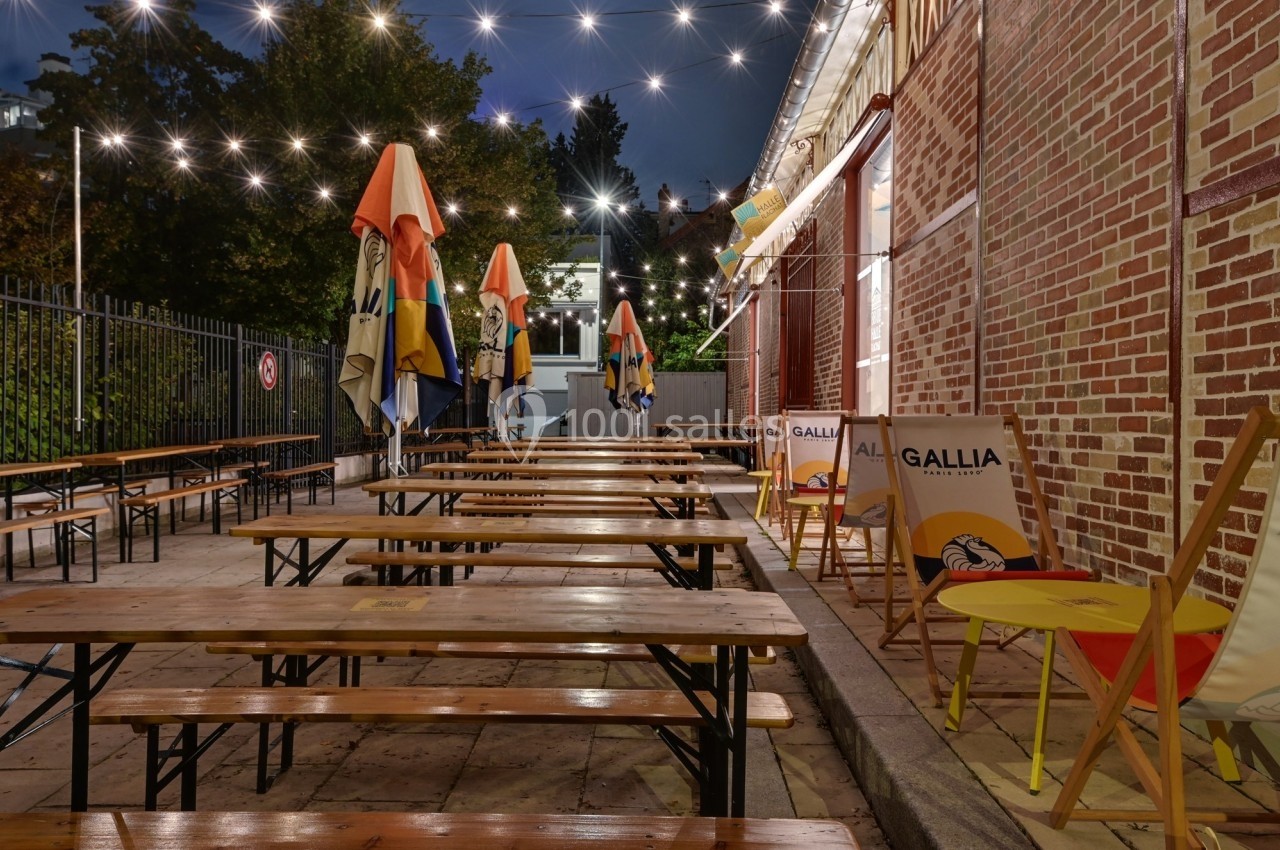 Terrasse extérieure avec tables en bois, chaises et parasols colorés, éclairée par des guirlandes lumineuses en soirée.
