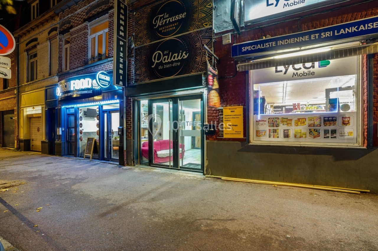 Façade de plusieurs commerces, dont un restaurant, une épicerie et une boutique de téléphonie, dans une rue éclairée.