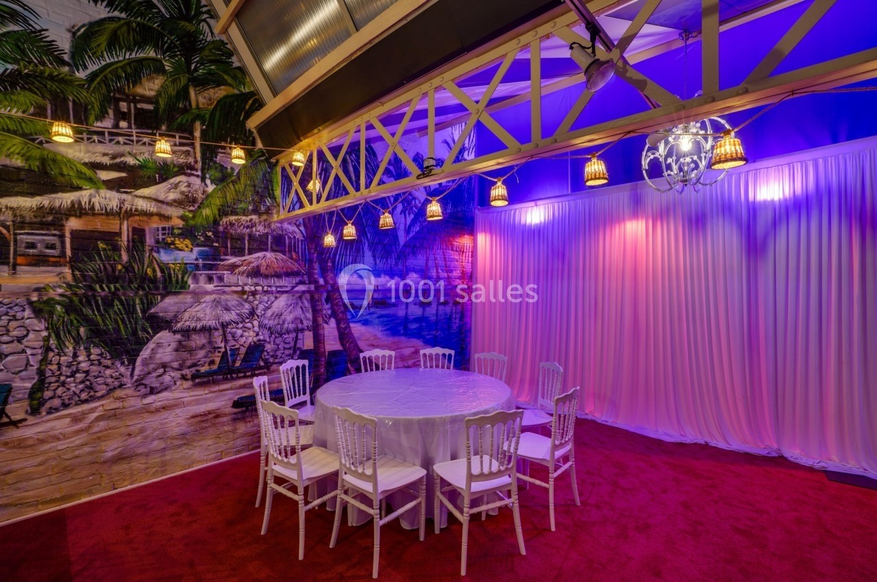 Salle décorée avec un mur représentant une plage, une table ronde entourée de chaises blanches et un éclairage coloré.