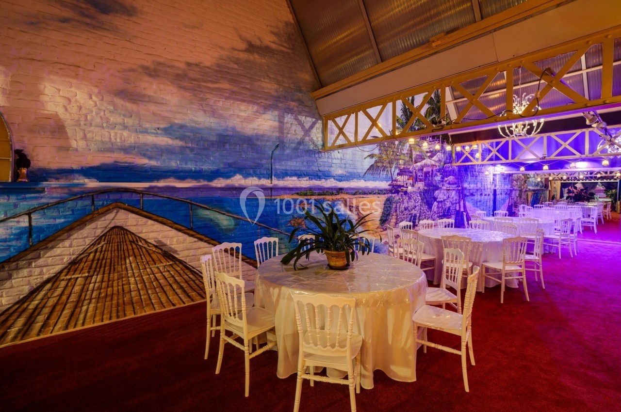 Salle décorée avec des tables rondes, chaises blanches et fresque murale représentant un paysage tropical.