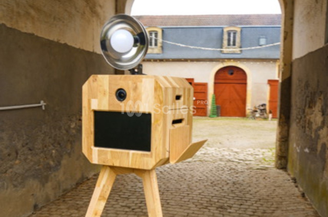 Boîte photo en bois sur trépied avec flash, placée dans une cour pavée devant des bâtiments anciens.