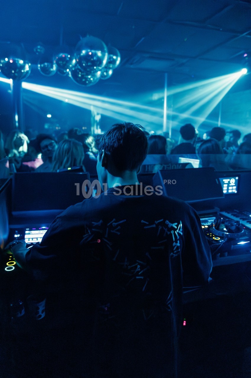 Un DJ mixe dans une boîte de nuit sombre, éclairée par des lumières bleues et des boules à facettes.