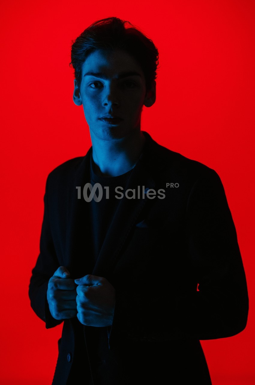 Un homme en costume sombre, éclairé par une lumière bleue sur fond rouge vif.