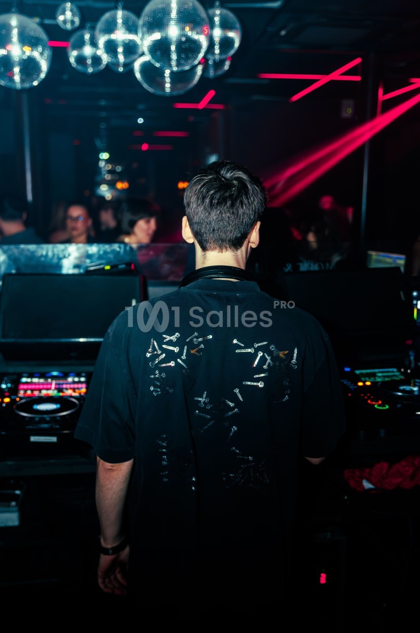 Un DJ de dos mixe dans une boîte de nuit sombre, éclairée par des lumières rouges et des boules à facettes.