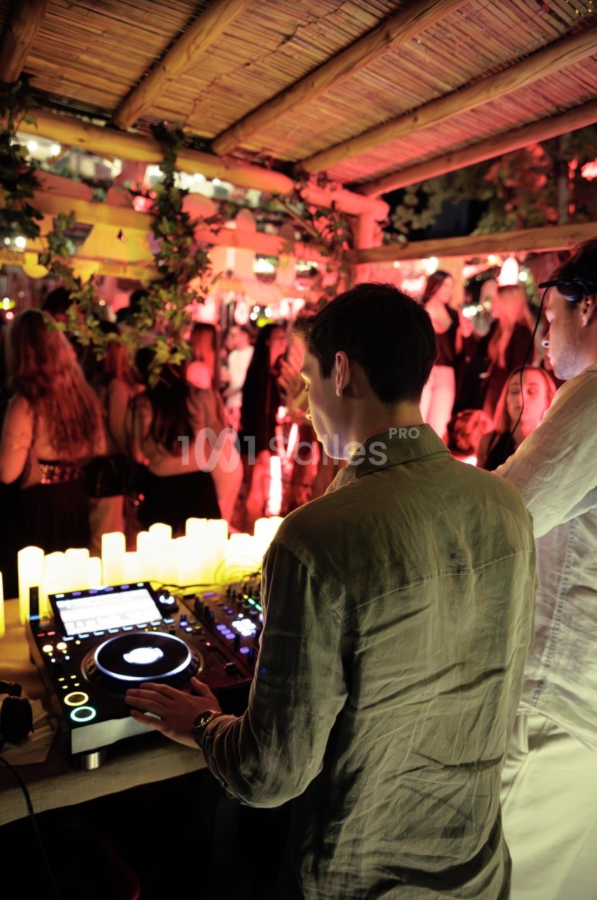 Un DJ mixe sur une console dans une ambiance festive avec des lumières tamisées et un public en arrière-plan.