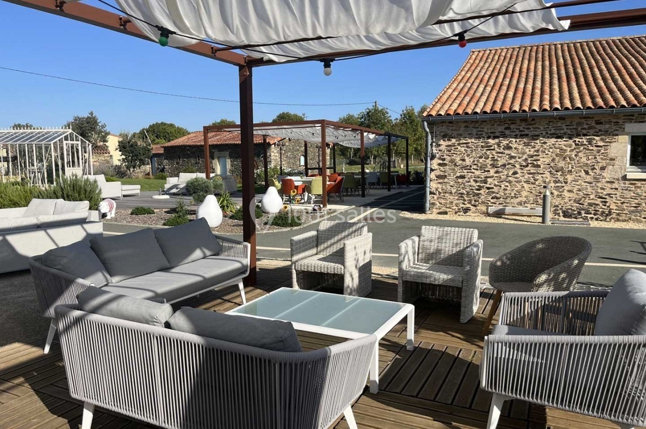 Terrasse aménagée avec des fauteuils et canapés en rotin, sous une pergola, près de bâtiments en pierre.