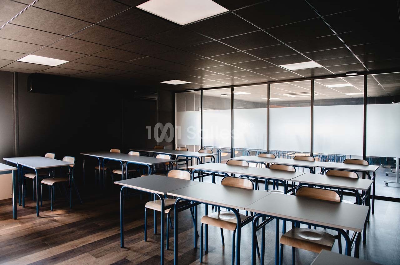 Salle de classe vide avec des tables et des chaises alignées, éclairée par des plafonniers et une lumière naturelle.