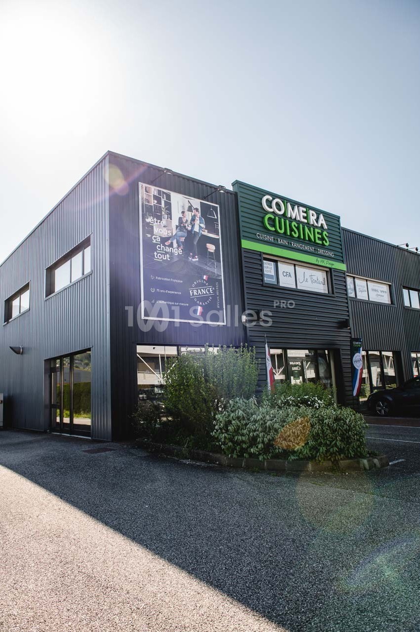 Façade d'un bâtiment commercial avec enseigne ’Comera Cuisines’ et affiches publicitaires, entouré de végétation.
