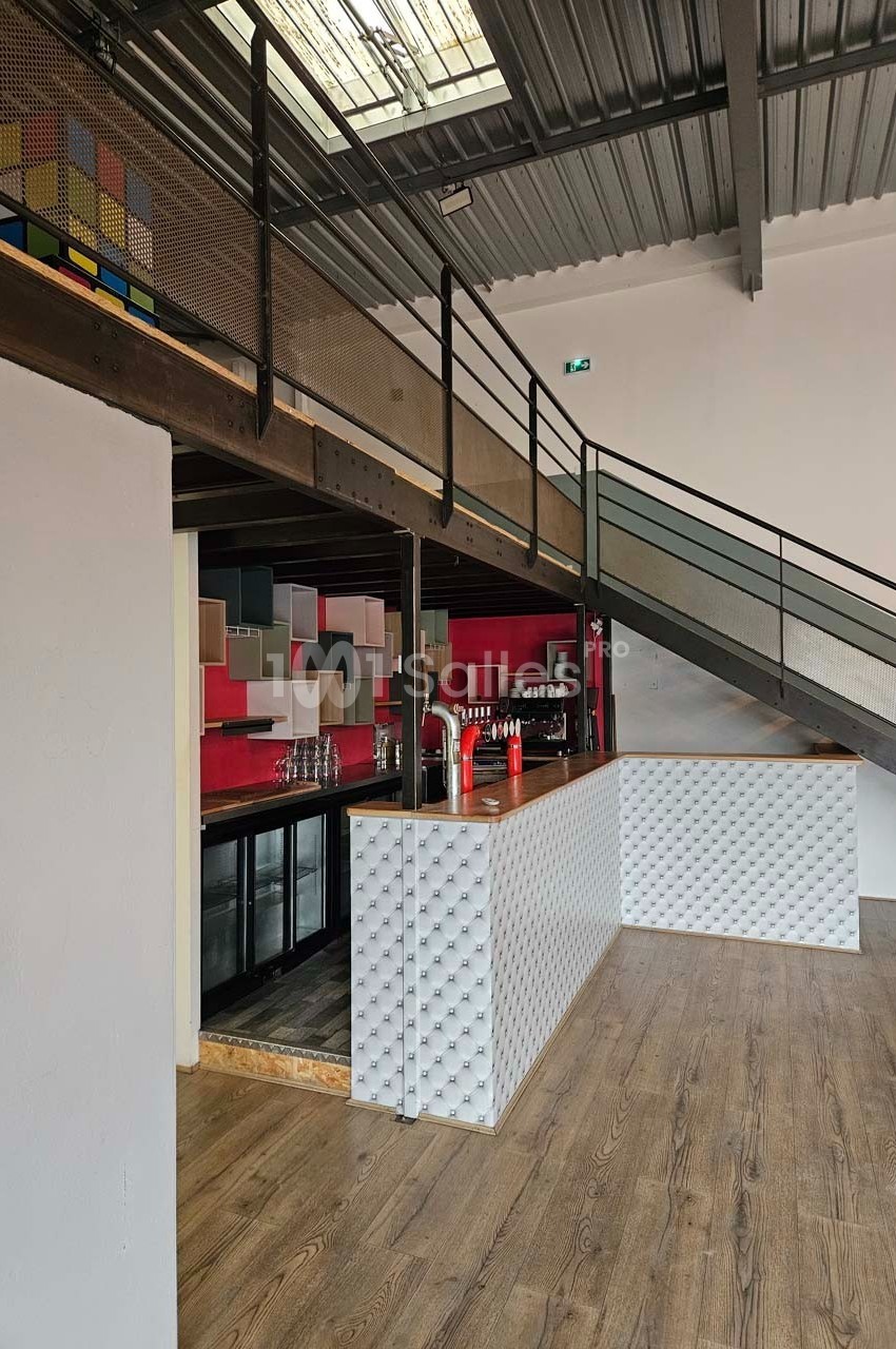 Un bar moderne avec comptoir texturé, sol en bois, mezzanine métallique et décoration colorée en arrière-plan.