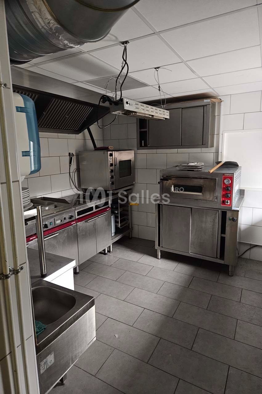 Cuisine professionnelle équipée avec four, plan de travail en inox, évier et hotte dans un espace carrelé.