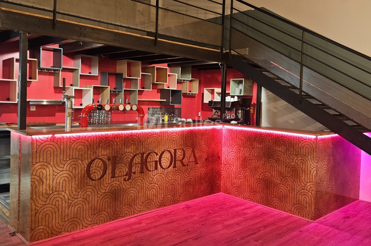 Bar moderne avec comptoir en bois gravé, étagères géométriques rouges et escalier métallique adjacent.