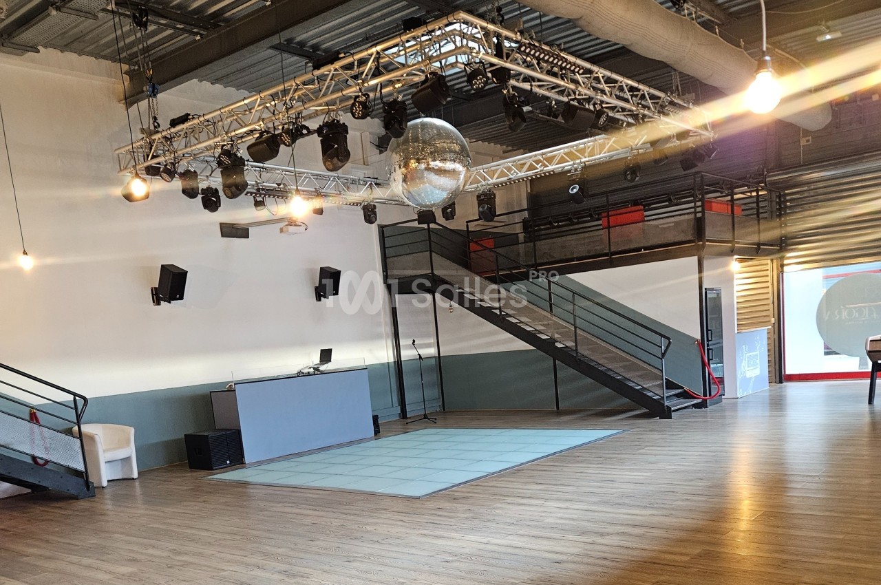 Salle spacieuse avec parquet, piste de danse, éclairage suspendu, boule disco et mezzanine accessible par un escalier.