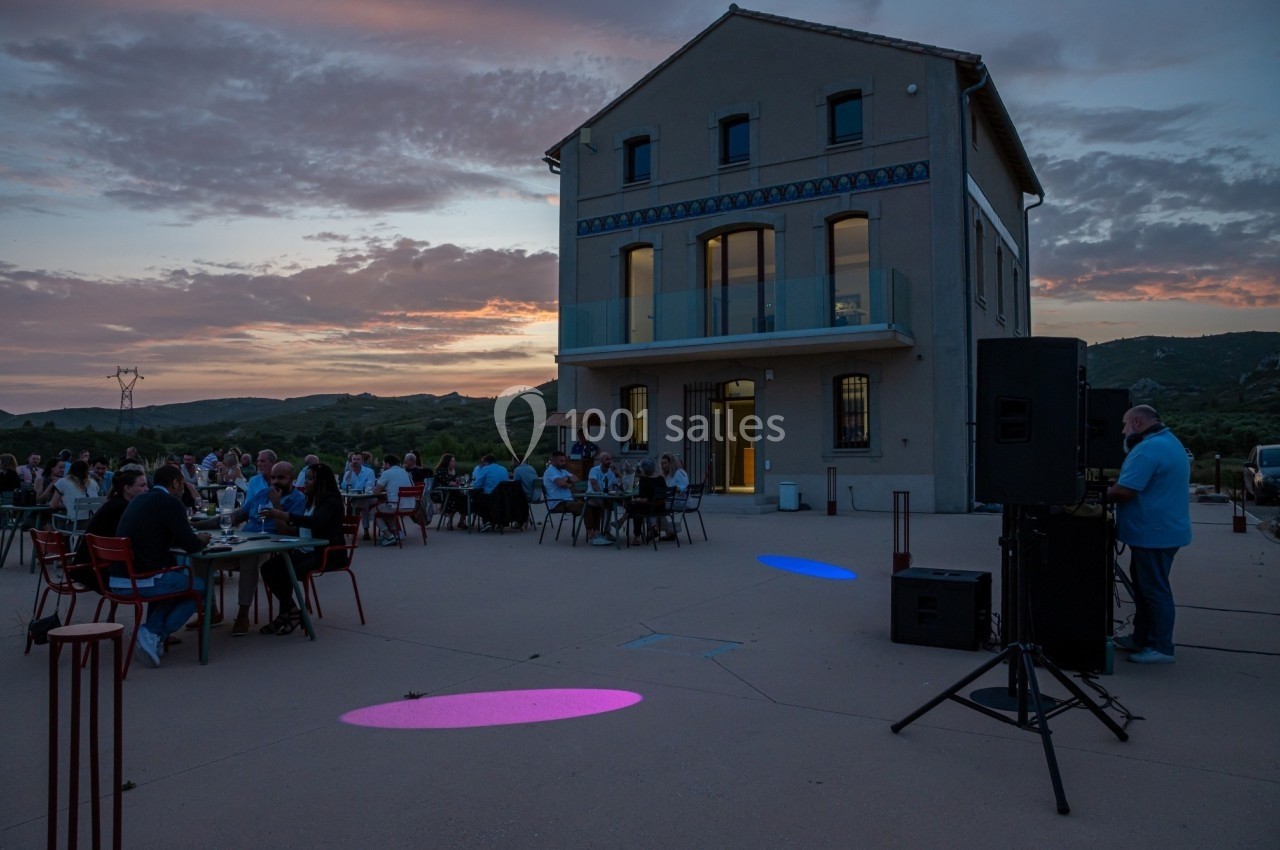 Groupe de personnes dînant en plein air au crépuscule devant un bâtiment moderne avec vue sur la campagne.