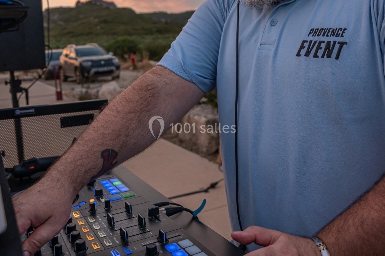 Un homme en polo bleu manipule une table de mixage en extérieur, avec une voiture et un paysage en arrière-plan.