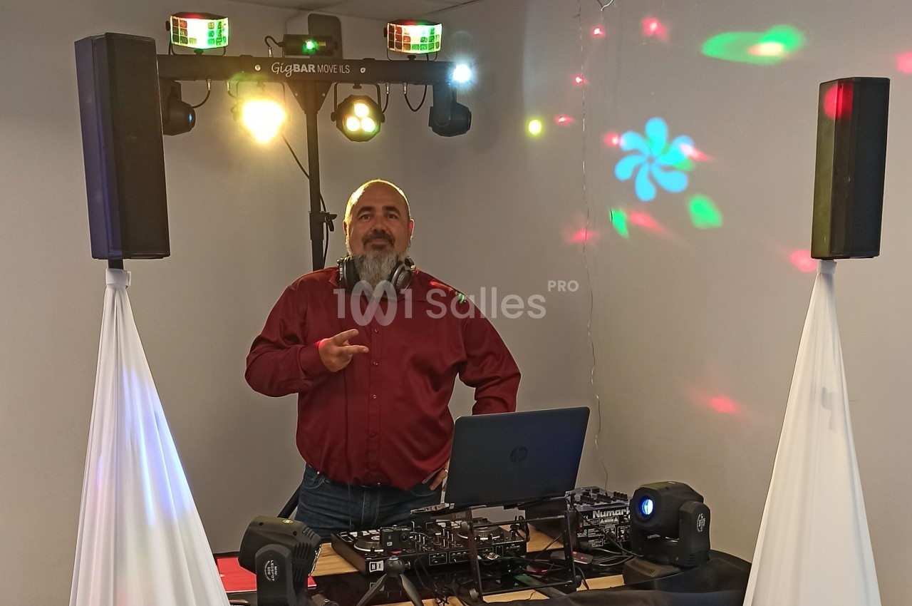 Un DJ en chemise rouge mixe de la musique sur une table équipée, avec des lumières et des haut-parleurs en arrière-plan.