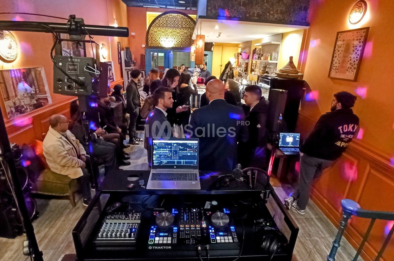 Vue d'une soirée dans un bar avec des invités debout, un DJ et des équipements de mixage au premier plan.