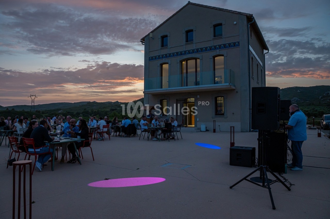 Groupe de personnes dînant en plein air au crépuscule devant un bâtiment moderne avec vue sur la campagne.