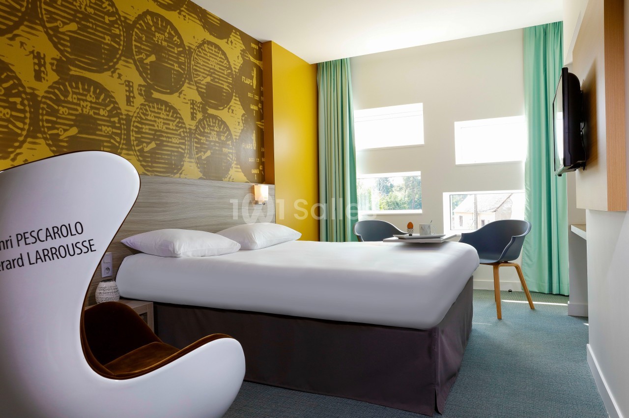 Chambre d'hôtel lumineuse avec un grand lit, fauteuil design, bureau, et fenêtres laissant entrer la lumière naturelle.