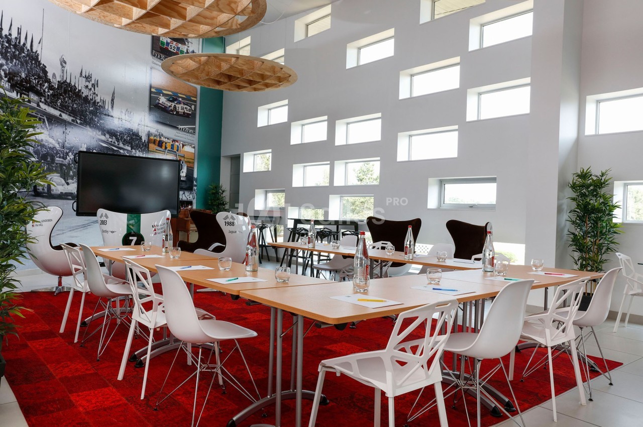 Salle de réunion lumineuse avec tables en bois, chaises blanches, écran, plantes et décoration murale.
