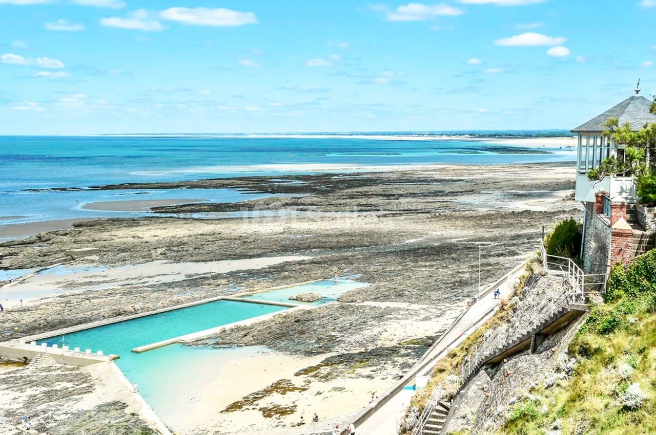 Vue d'une piscine d'eau de mer surplombant une plage rocheuse et l'océan sous un ciel bleu dégagé.