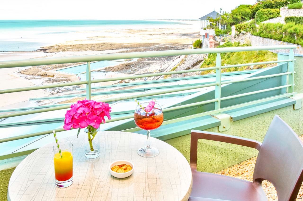 Vue sur une plage depuis une terrasse avec table, boissons, fleurs roses et fauteuils, entourée de verdure.